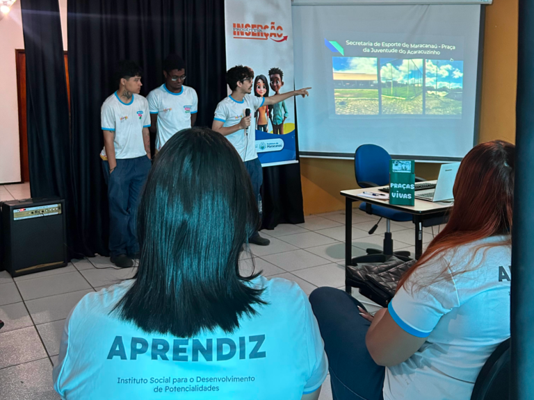 Jovens aprendizes do idep, vestindo camisetas do programa Aprendiz, apresentam um projeto sobre inserção produtiva para uma plateia sentada.